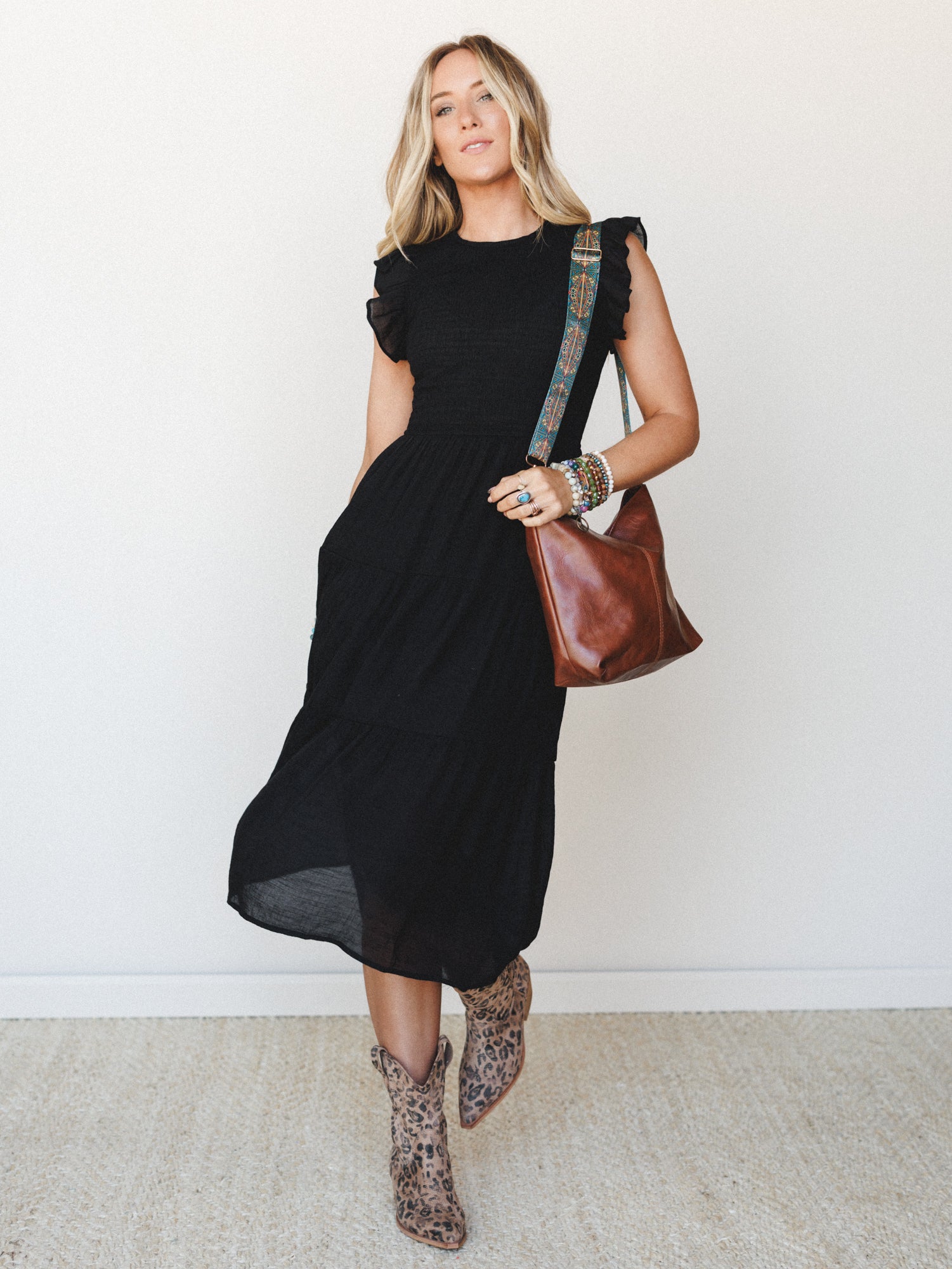 A black midi dress with flutter sleeves and a tiered skirt styled with leopard print cowboy boots, a cognac leather crossbody bag with a boho embroidered strap, and stacked beaded bracelets — the kind of outfit that works just as hard at a concert as