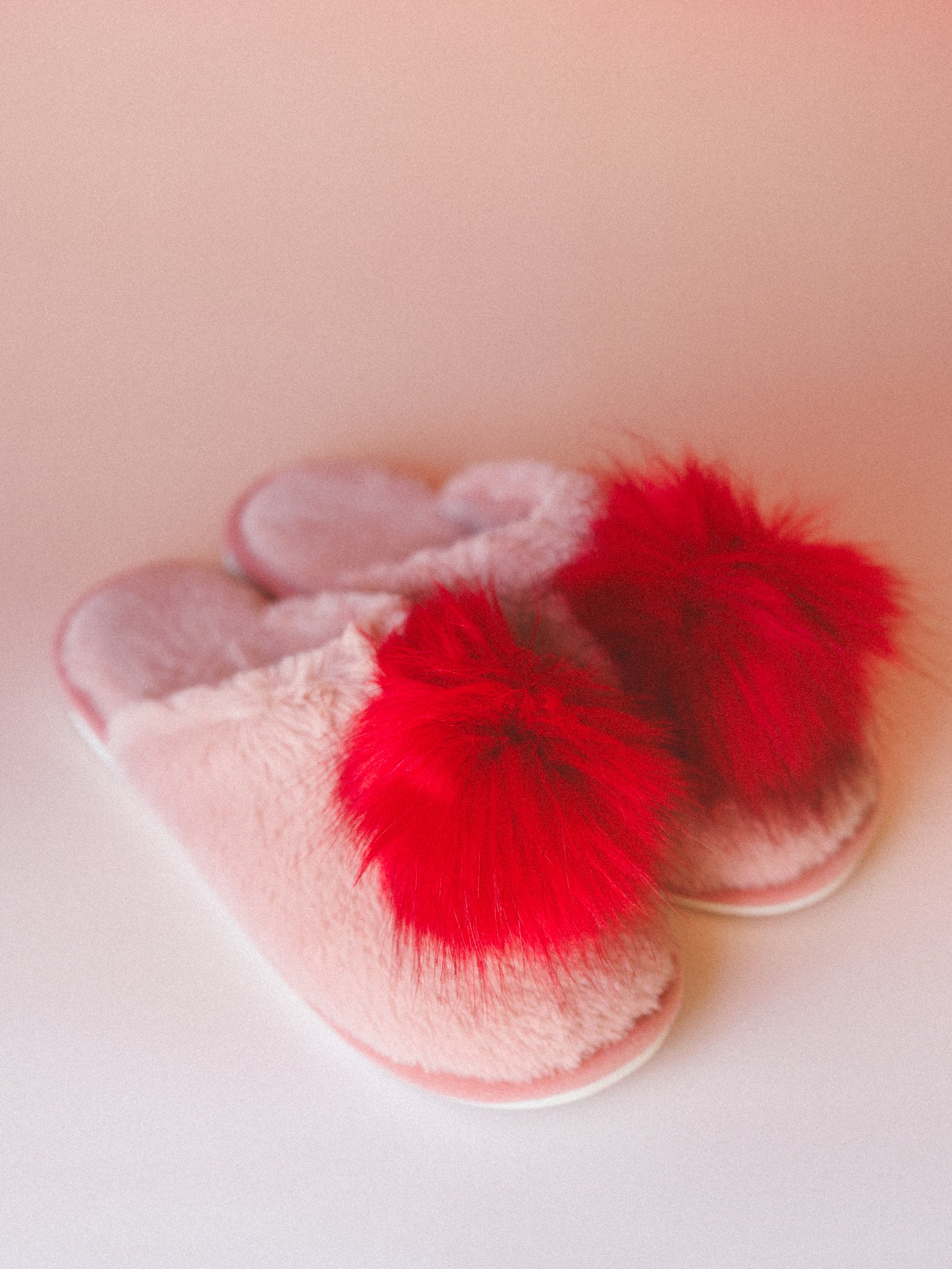 A pair of blush pink fuzzy slide slippers with bold red pom-pom trim, shot on a soft pink surface — the kind of thing that makes staying in feel intentional.