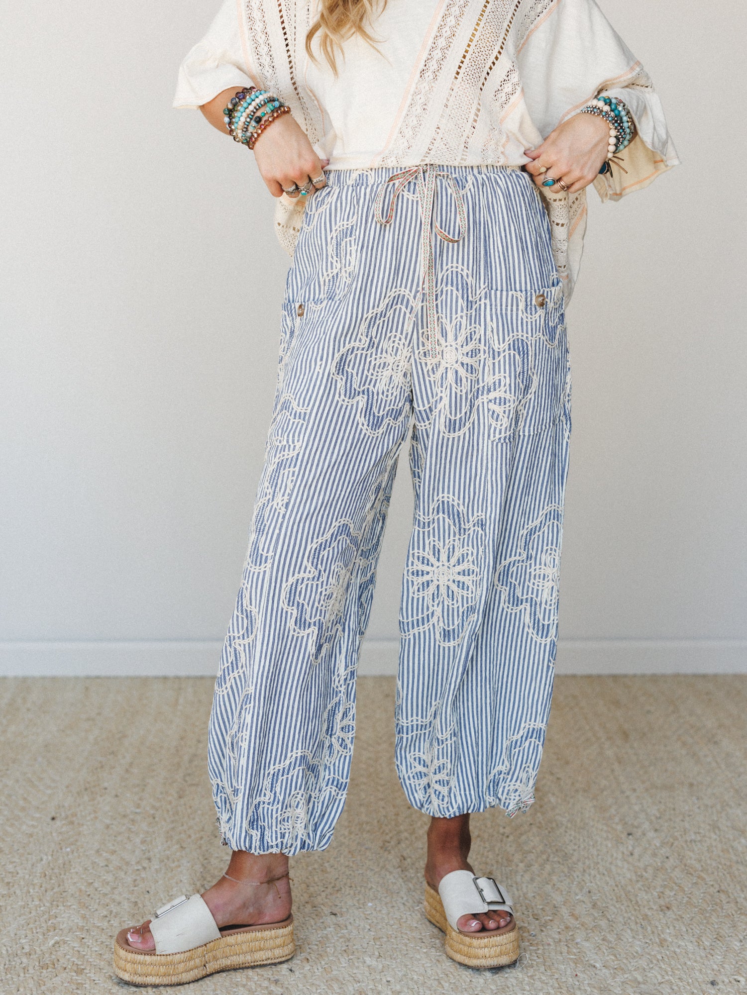 A breezy mid-shot of wide-leg striped pants in denim blue and cream, covered in rope-style floral embroidery, styled with a cream lace top, layered bead bracelets, and white platform espadrilles — the kind of outfit that feels equally at home at a co