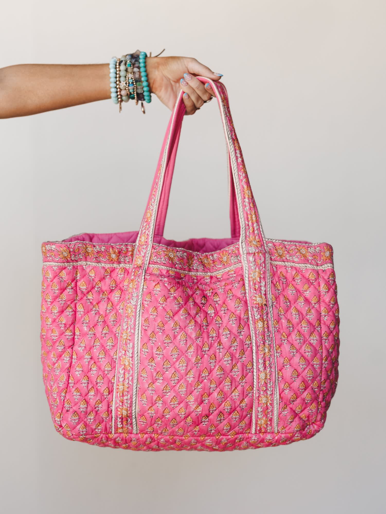 A hand holds up a roomy quilted tote in bubbly pink with a block-print floral motif, styled with a stack of beaded boho bracelets — the kind of bag that goes everywhere and makes it look intentional.