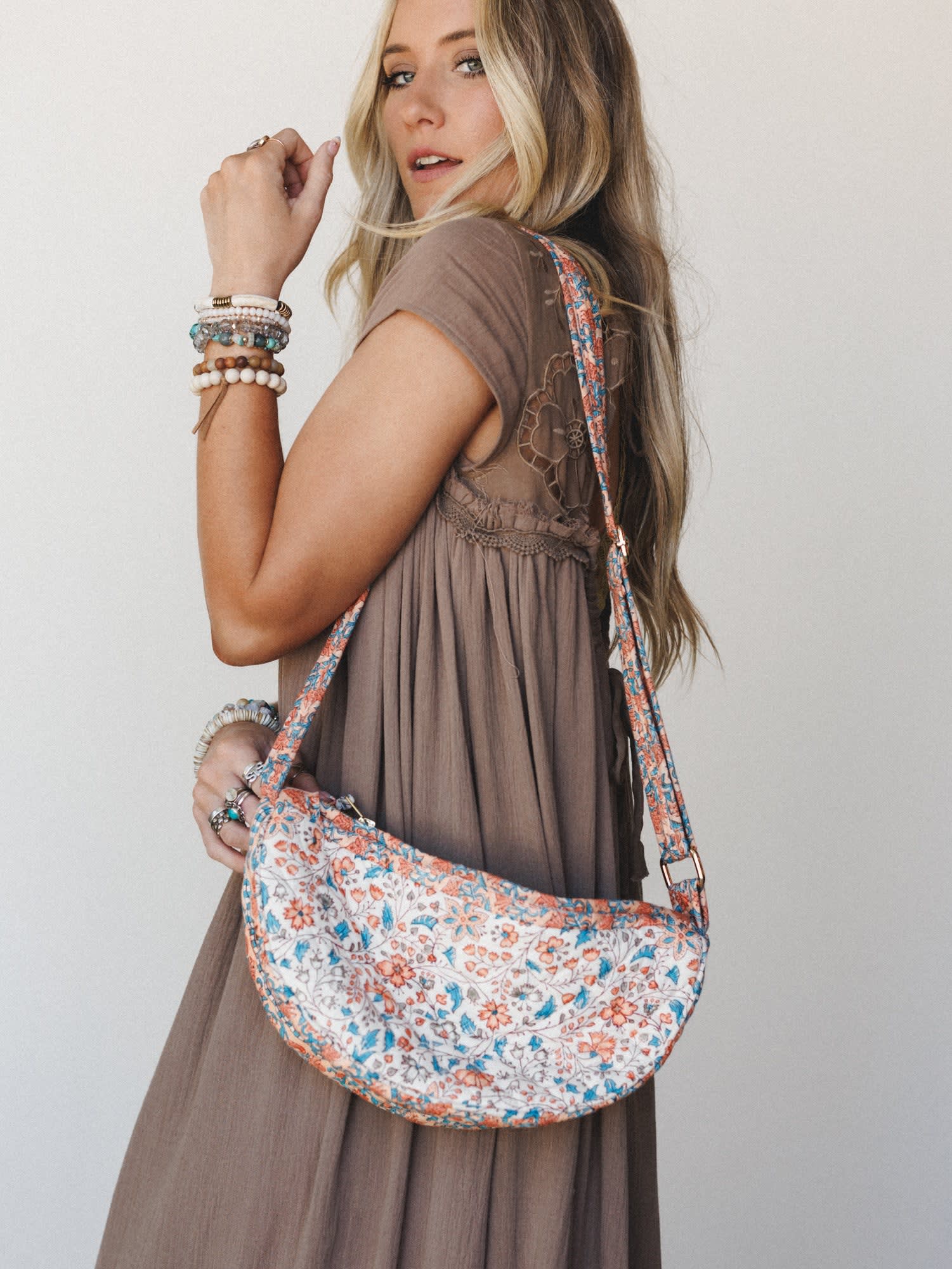 A sun-kissed model glances back over her shoulder wearing a flowy taupe maxi dress with lace back detail, styled with a crescent-shaped floral quilted crossbody bag and layers of mixed beaded bracelets — the whole look feels like a good Saturday with
