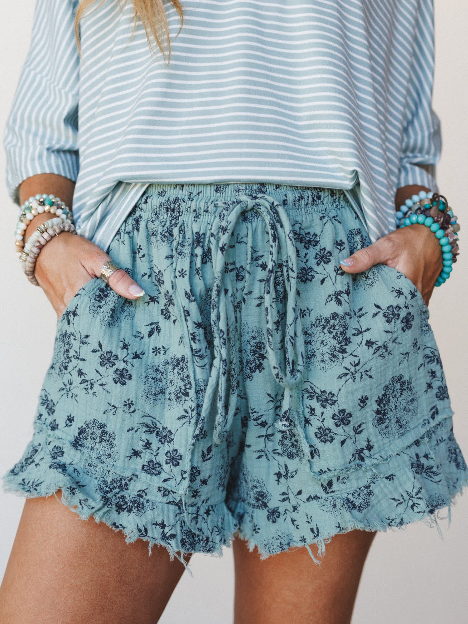 A close-up mid-shot of flowy floral shorts in dusty turquoise paired with a blue stripe top — hands tucked into oversized patch pockets, stacked bracelets on both wrists, the whole thing feeling like a lazy summer morning that somehow looks this good