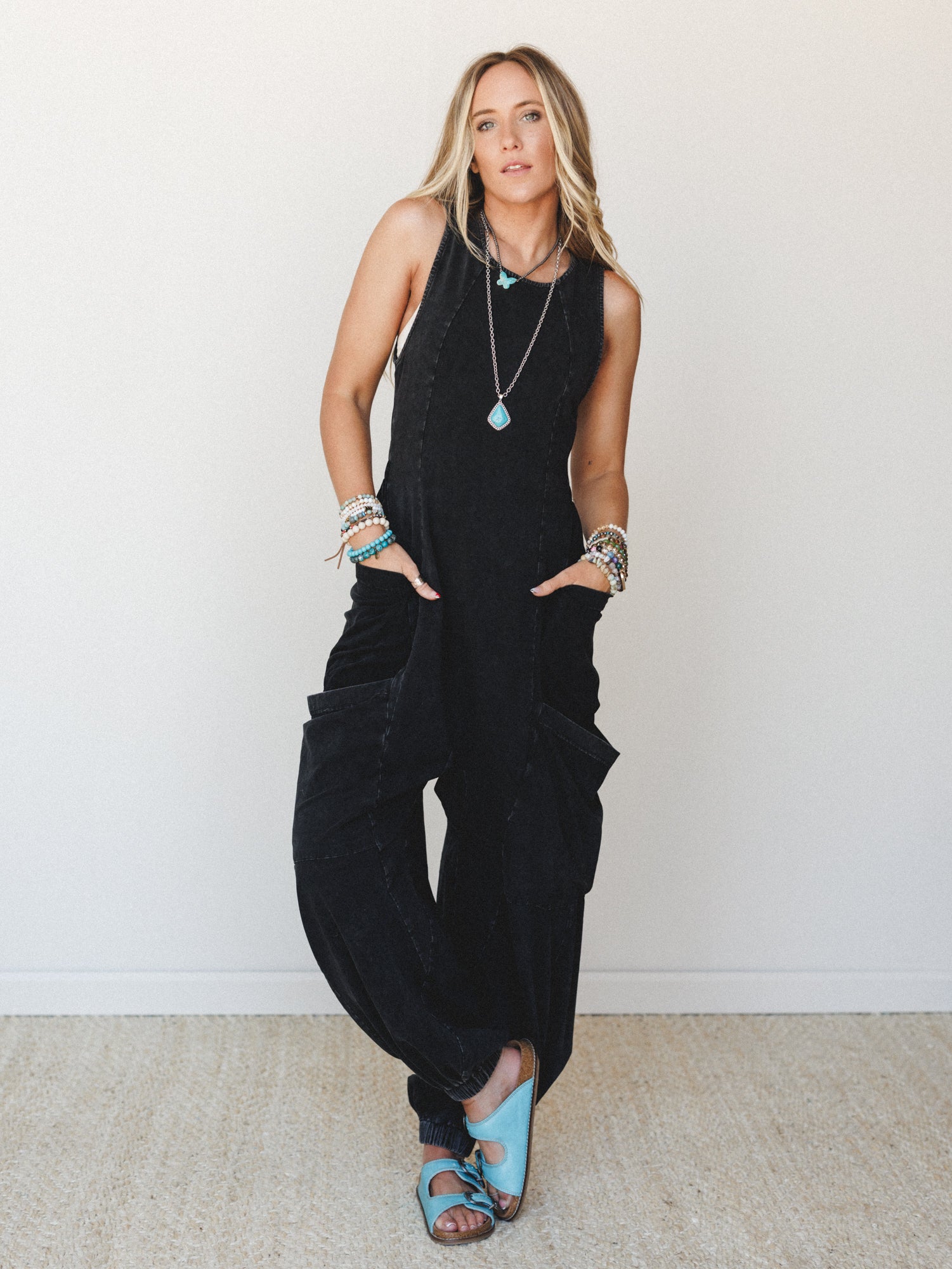 A relaxed, washed black cargo jumpsuit styled with turquoise sandals and layered boho jewelry — the kind of effortless one-and-done outfit that looks like you tried just enough.