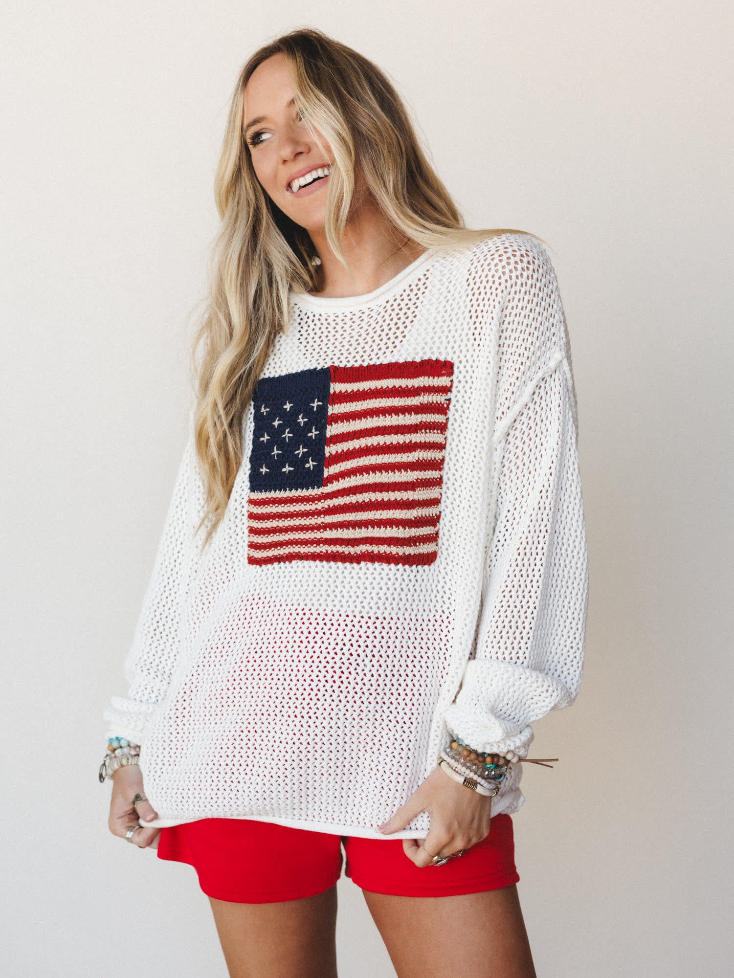 A smiling model in an oversized ivory open-knit sweater with a bold American flag intarsia graphic paired with red shorts and stacked boho bracelets — the kind of effortless Fourth of July outfit that looks like it took zero effort and exactly the ri