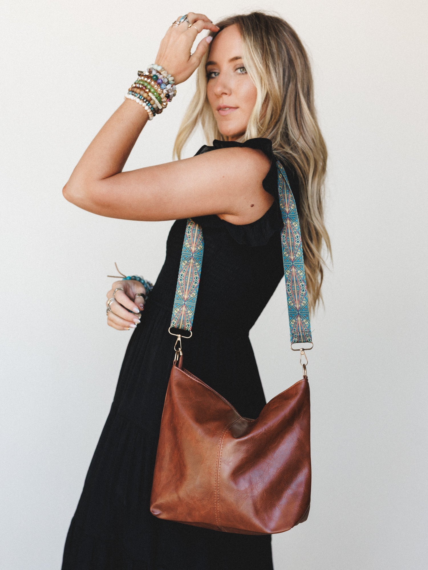 A model in a sleeveless black dress poses in three-quarter profile, showing off a slouchy camel leather hobo bag with a boho geometric printed strap — stacked beaded bracelets and rings layered on, the whole look effortlessly personal.