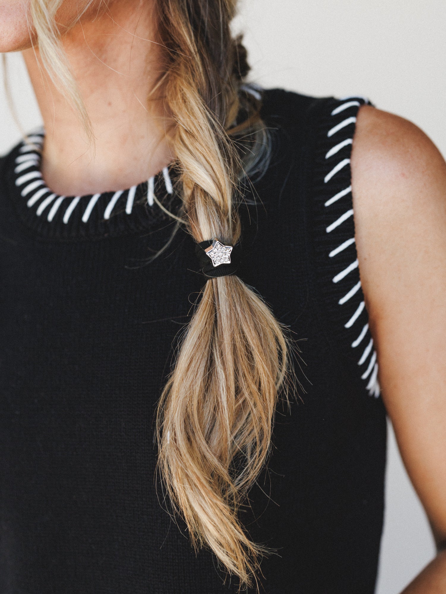 A close-up lifestyle shot highlighting a rhinestone star hair tie holding a loose, effortless braid against a black sleeveless sweater vest — the kind of little detail that quietly makes the whole look.