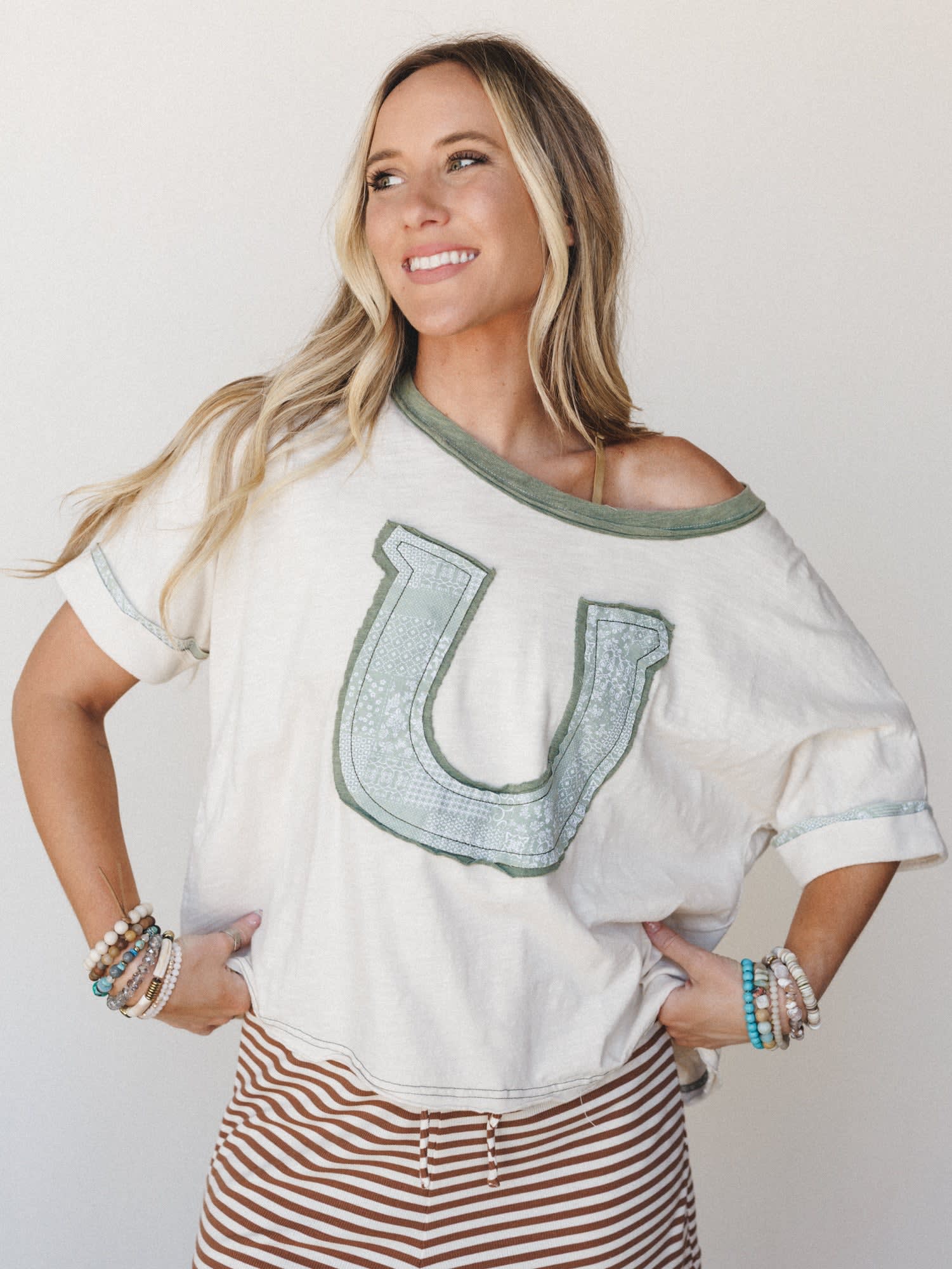 A relaxed, off-shoulder graphic tee in cream with a sage green horseshoe appliqué patch takes center stage, styled with rust-striped bottoms and stacked boho bracelets — the kind of effortless outfit that feels as good as it looks.