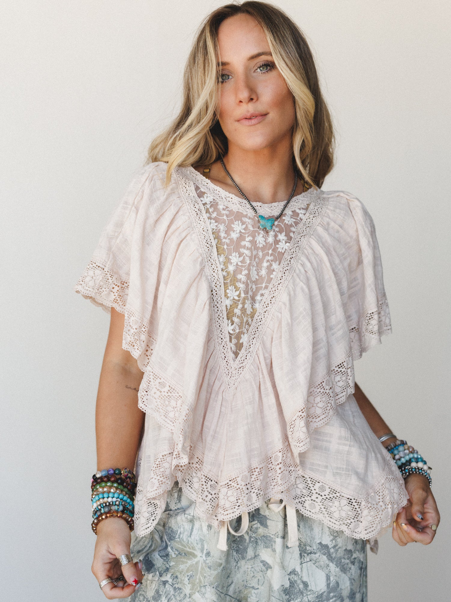 A flowy blush lace-trimmed top with an embroidered sheer V-neckline panel sits softly on the model, styled with stacked boho bracelets and a turquoise pendant — the kind of top that looks like it was made for slow mornings and good light.