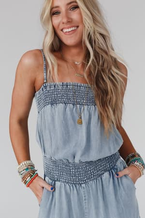 A woman with long, wavy blonde hair smiles at the camera in the Harlow Wide Leg Chambray Jumpsuit - Blue, layered necklaces, and colorful beaded bracelets, her hands in her pockets against a plain background.