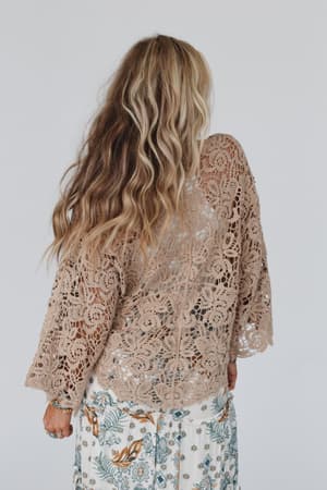 A woman with long, wavy blonde and brown hair stands facing away, wearing the Crochet Petal Kimono Wrap - Mocha over a white skirt with blue florals—a chic boho piece—set against a simple light background.