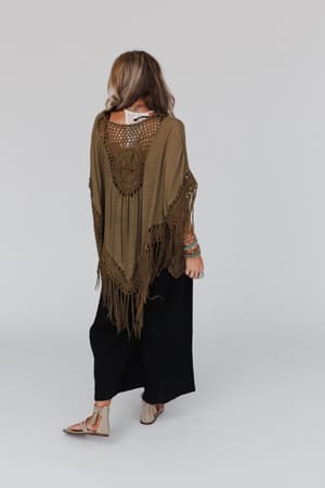 A woman faces away, wearing the Dreamy Macramé Kimono in olive over a white top and black wide-leg pants. She has light brown hair, sandals, colorful bracelets, and stands against a plain light background.