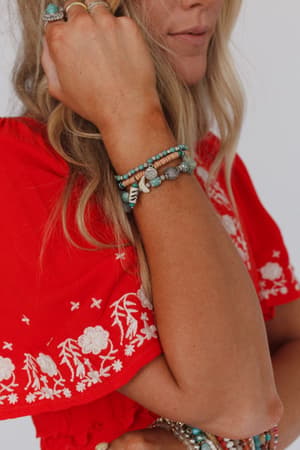 A woman in a red embroidered top shows off her wavy blonde hair and the San Antonio Bracelet Set - Turquoise. Her raised arm, adorned with these boho beaded bracelets, partially covers her face.