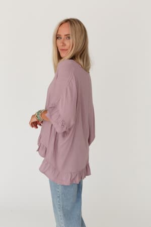A woman with long blonde hair wears The Nest Let Me Be Ruffled Top in Pale Plum, featuring a lightweight stretch knit, v-neckline, and loose sleeves. She pairs it with light blue jeans and smiles slightly against a plain white background.
