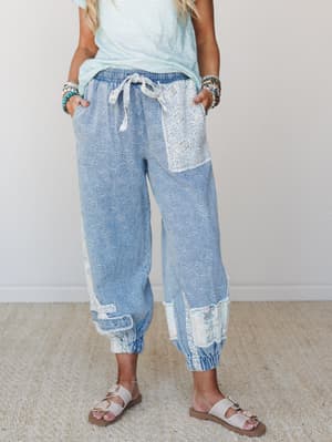 A relaxed pair of acid-washed denim joggers with floral embroidery and lace patchwork details, styled with a mint tee, stacked bracelets, and sandals — the kind of pants that feel like a whole personality decision.