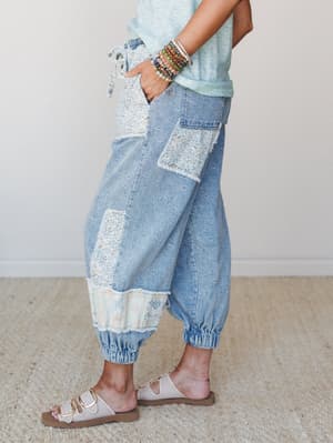 A side-profile shot of patchwork denim joggers styled with a soft mint tee, stacked bead bracelets, and slide sandals — the kind of pants that somehow look intentional whether you're running errands or at a weekend market.