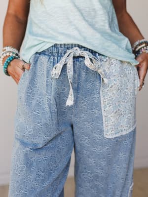 A close-up of relaxed wide-leg denim pants with an allover floral burnout texture, lace drawstring tie, and a contrast floral fabric pocket insert — styled with a mint slub tank and stacked beaded bracelets for an effortlessly boho everyday look.