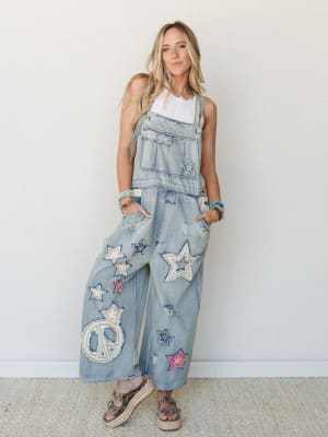 Wide-leg light denim overalls covered in star and peace sign appliqué patches styled over a white lace tank, paired with platform espadrille sandals and stacked bead bracelets — the kind of outfit that makes a whole room turn around.