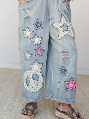 A close-up of wide-leg light wash denim overalls covered in frayed star appliqués and a peace sign patch — the kind of pants that do all the talking so you don't have to.