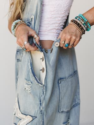 A close-up detail shot of light wash distressed denim overalls with star patchwork and button fly, styled with a soft pink textured crop top and stacked with turquoise, wood bead, and silver jewelry — the kind of outfit that's clearly been lived in a