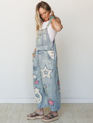 Light-wash denim overalls covered in star patches and a peace sign appliqué, styled over a white lace tank with stacked turquoise bracelets and leopard platform sandals — playful, free-spirited, and a little bit nostalgic in the best way.