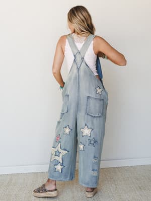 Shot from the back, a model in wide-leg light denim overalls covered in mixed star appliqués pairs them with a white lace tank and espadrille sandals — the kind of outfit that makes a regular Tuesday feel like a road trip.