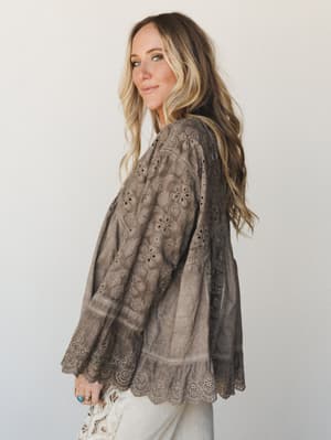 A flowy eyelet blouse in a warm mushroom brown, shot from a side angle — the tiered silhouette and scalloped hem do all the talking while she glances back over her shoulder like she already knows it's a good outfit.