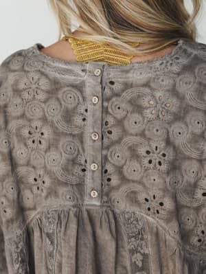 A close-up back detail shot of a dusty mushroom-toned eyelet blouse, showing intricate floral and swirl embroidery, a center back button placket, gathered waist seam, and a peek of a mustard lace hair accessory at the nape — all the quiet details tha