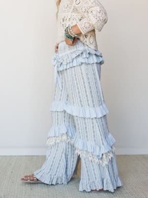 A dreamy side-profile shot of wide-leg pants in a soft blue floral stripe, layered with tiered ruffles and lace trim, styled with a cream crochet top and pearl sandals — the kind of outfit that makes a regular afternoon feel a little more like someth