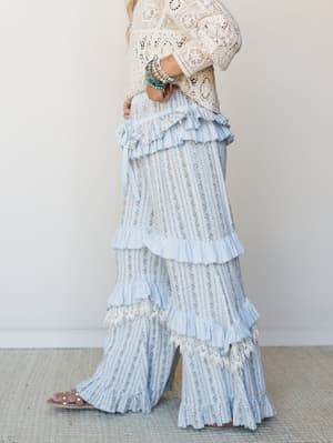 A dreamy side-profile shot of wide-leg pants in a soft blue floral stripe, layered with tiered ruffles and lace trim, styled with a cream crochet top and pearl sandals — the kind of outfit that makes a regular afternoon feel a little more like someth