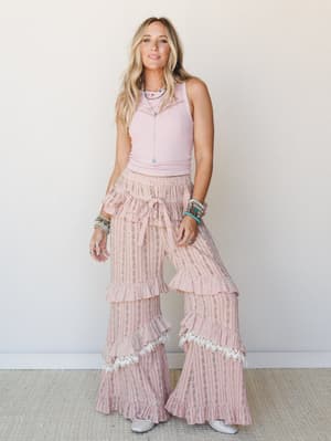 A blonde model in a bright studio setting wears wide-leg ruffle bell pants in a soft peach floral stripe, paired with a ribbed pink crop tank and stacked turquoise jewelry — the whole look has that sweet, free-spirited energy that lives somewhere bet