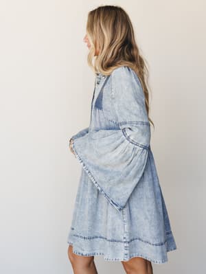 A light acid-wash denim mini dress with dramatic bell sleeves and pintuck detailing shot in profile against a soft studio backdrop — the kind of dress that looks equally at home at a farmers market or a music festival.