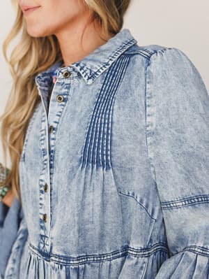 A close-up detail shot of a light acid-wash denim dress with pintuck pleating down the bodice, brass snap buttons, and a gathered tiered skirt — the kind of piece that looks like it was made for wandering somewhere with good light.