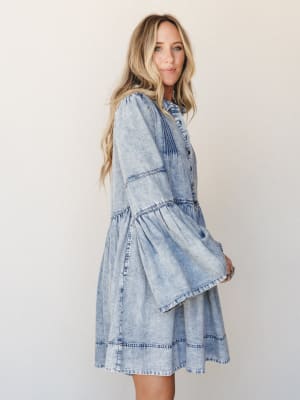 A light acid-wash denim dress with tiered skirt and dramatic bell sleeves photographed against a clean studio wall — effortlessly boho with a vintage western edge.