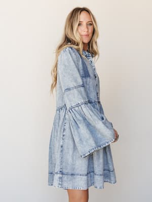 A light acid-wash denim dress with tiered skirt and dramatic bell sleeves photographed against a clean studio wall — effortlessly boho with a vintage western edge.