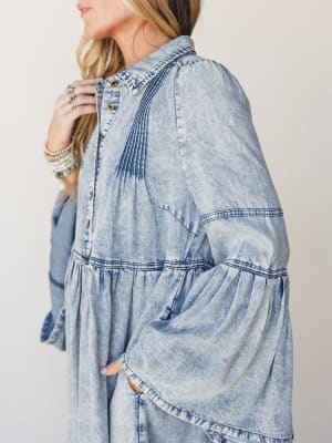 A close-up side shot of a flowy acid-wash denim dress with dramatic bell sleeves and pintuck detailing — styled with stacked turquoise and pearl bracelets for that effortless boho-western feel.