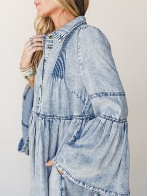 A close-up side shot of a flowy acid-wash denim dress with dramatic bell sleeves and pintuck detailing — styled with stacked turquoise and pearl bracelets for that effortless boho-western feel.