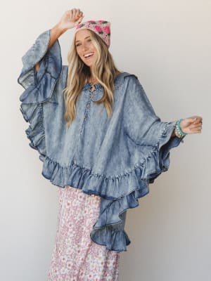 A smiling model wears an oversized acid-wash denim ruffle poncho over a pink floral maxi skirt, topped with a pink crochet strawberry beanie — the whole look reads like a boho dream you'd actually get dressed in.