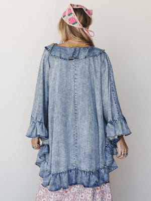 Shot from behind, a model wears a flowy acid-wash denim poncho with ruffled trim at the neckline and tiered bell sleeves, layered over a floral skirt and finished with a pink crochet headscarf — the whole look has that effortless, slightly dreamy ene