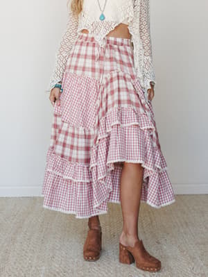 A tiered high-low maxi skirt in mixed pink plaid and gingham is styled with a cropped open-knit crochet top, wood-heeled clogs, and turquoise jewelry — the kind of outfit that feels like a farmers market and a country concert at the same time.