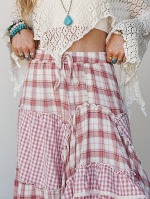 A close-up of a mixed plaid and gingham tiered maxi skirt in dusty rose and cream, paired with a cropped crochet top and layered turquoise jewelry — the kind of outfit that feels like a summer afternoon that got away from you in the best way.