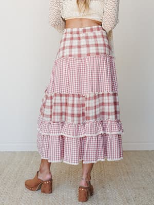 Shot from behind, a tiered plaid maxi skirt in rosy pink and cream pairs with an open-knit cropped top and wooden platform clogs — the kind of outfit that feels like a farmers market and a good playlist all at once.