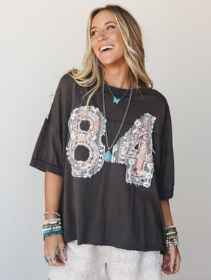 An oversized washed black graphic tee with lace-and-floral patchwork '84' appliqué, styled with white eyelet bottoms and layers of turquoise and beaded jewelry — the kind of top that looks like it's been yours forever.