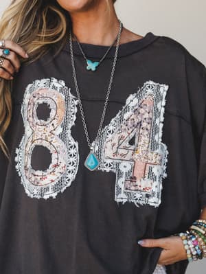 A washed black oversized graphic tee with floral and lace patchwork '84' appliqué, styled with layered turquoise necklaces, stacked beaded bracelets, and silver rings — the kind of top that looks like it has a whole personality before you even put it