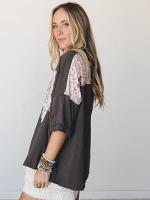 A relaxed, lived-in oversized tee in washed charcoal with floral patchwork paneling across the back and shoulders, styled with a white eyelet skirt and a stack of beaded bracelets — effortlessly boho without trying too hard.