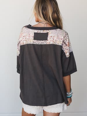 Shot from the back, this oversized washed-black tee does something unexpected — a patchwork floral yoke across the shoulders adds the kind of detail you notice when someone walks away, styled casually with lace-trim shorts and stacked turquoise brace