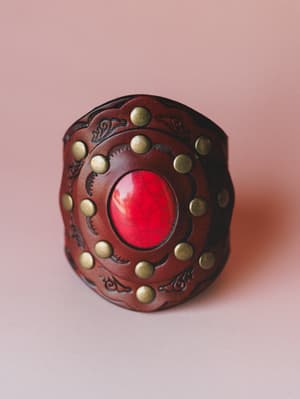 A wide leather cuff bracelet shot close-up against a dusty rose background — rich brown tooled leather with brass studs circling a bold red stone center, the kind of piece that looks like it has a story.