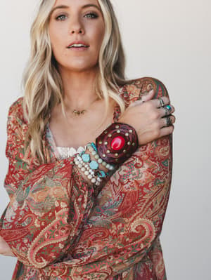 A close-up lifestyle shot of a model in a richly patterned paisley kimono, wrist stacked with a bold red stone leather cuff, beaded bracelets, and layered rings — the kind of arm party that looks collected, not coordinated.
