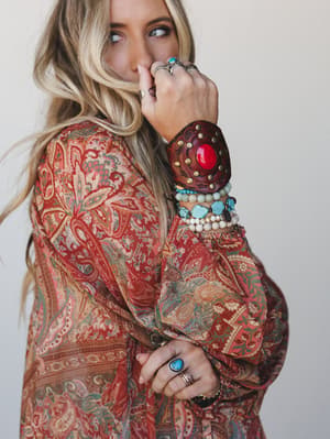 A close-up lifestyle shot highlighting a stacked arm party — leather cuff with a red stone center, beaded bracelets in turquoise and cream, and silver rings layered over a paisley kimono in warm rust and sage tones.