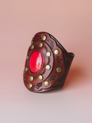 A wide leather cuff bracelet shot close-up against a blush studio backdrop — deep mahogany leather tooled with scrolling floral engravings, ringed with antique brass studs, and centered with a bold oval red cabochon stone that gives it an almost medi