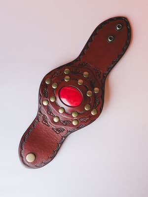 A wide leather cuff bracelet in deep oxblood brown, laid flat against a soft blush background — handcrafted-looking with tooled floral engravings, a circle of antique brass studs, and a glossy red cabochon stone at the center.